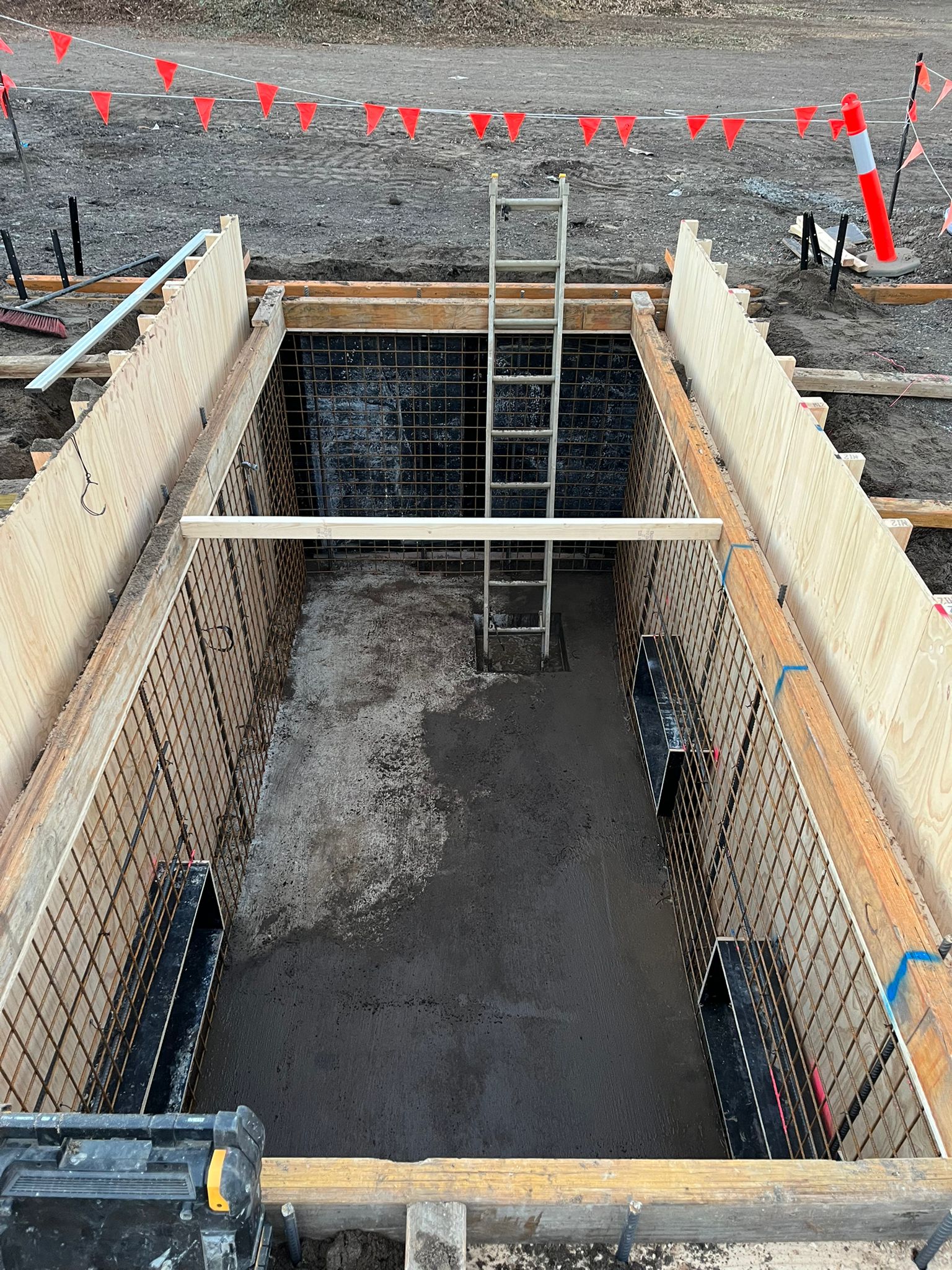 A rectangular concrete pit with a ladder inside, surrounded by wooden supports and safety flags.