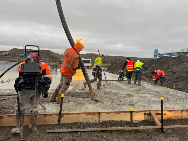 Construction workers in safety gear pour concrete onto a large roadway under cloudy skies.