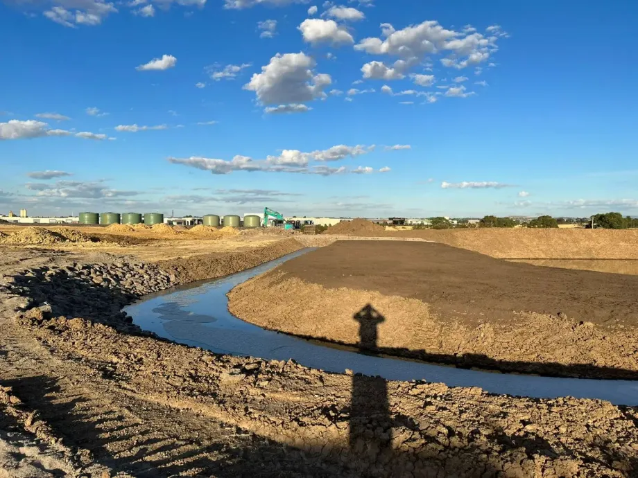 Excavated construction site with a small stream, clear sky, and machinery in the background.