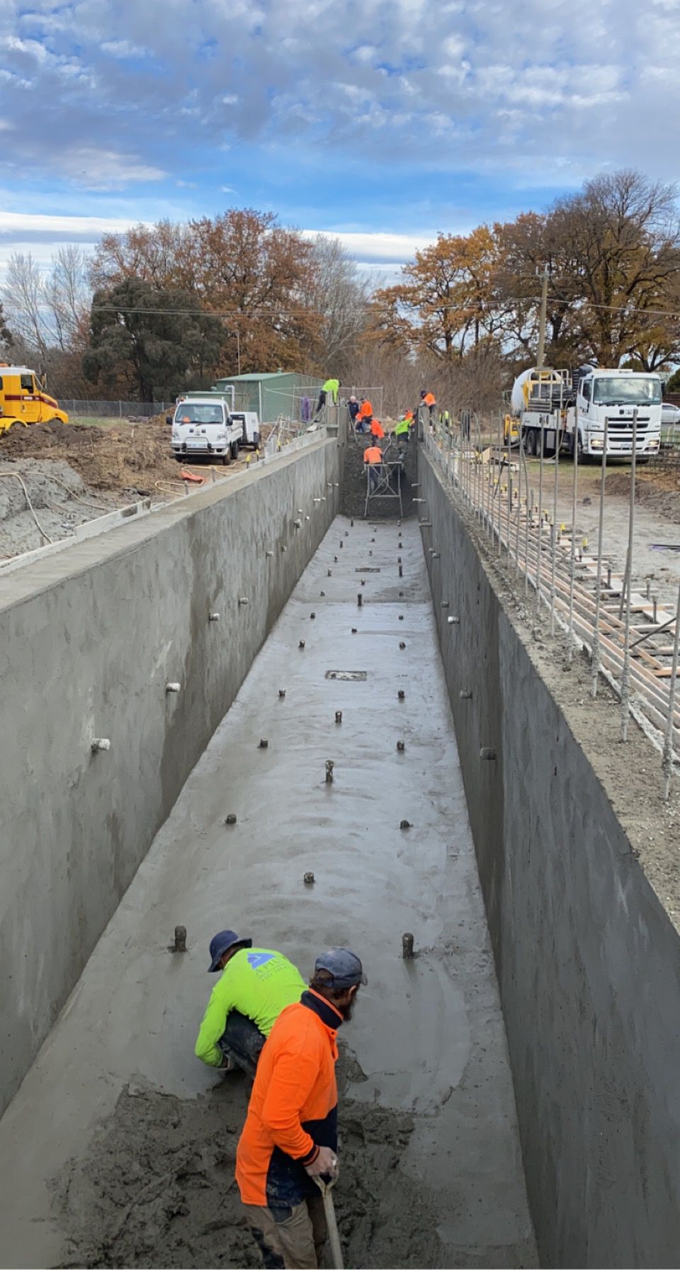Workers in safety gear smooth concrete in a large drainage channel beside parked construction vehicles.
