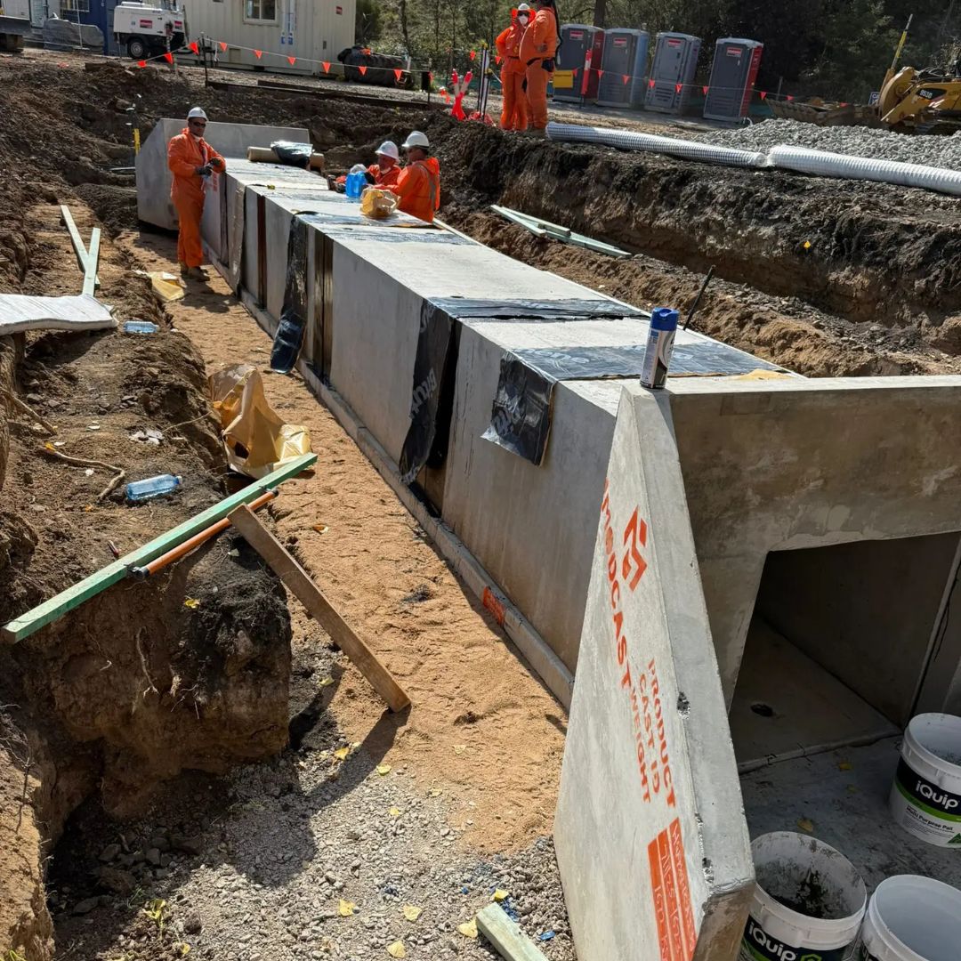 Workers in orange uniforms install large concrete sections in a trench at a construction site.