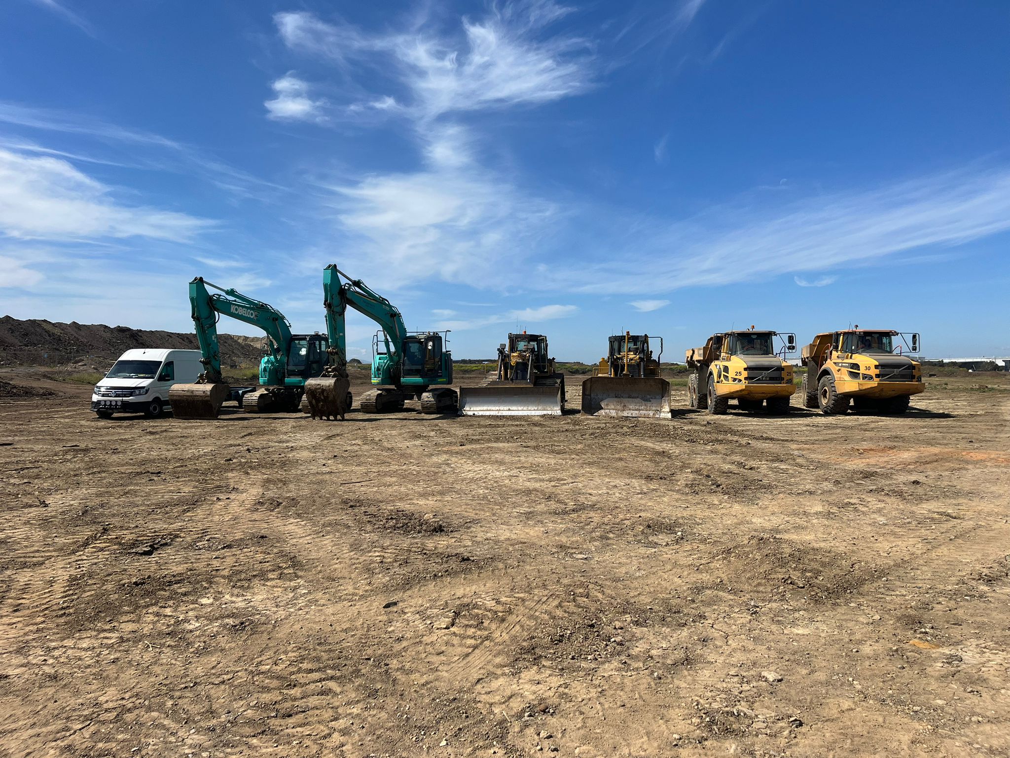 Six construction vehicles, including diggers and loaders, are lined up on a dirt field under a clear sky.