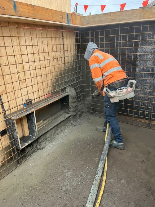 Worker in orange safety gear spraying concrete onto a rebar wall at a construction site.