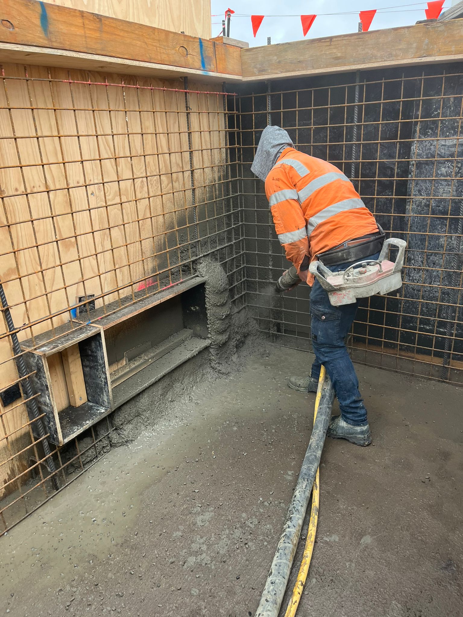 Worker in orange safety gear spraying concrete onto a rebar wall at a construction site.